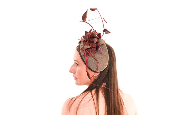 Glimmering Wine Percher is a beautiful classic vintage style cocktail hat with added height of feather vine and leather flower, hand made and wired on a buckram base covered with a fantastic sparkle fabric that is both silver or gold in different light. Model is staring off to the left and left shoulder towards the camera