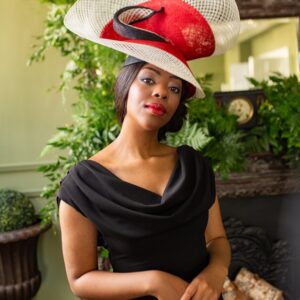 Model with brown skin and dark hair wears a red , ivory and black swirly freeformed hat. Large and bold with a cowl neck black fitted dress.